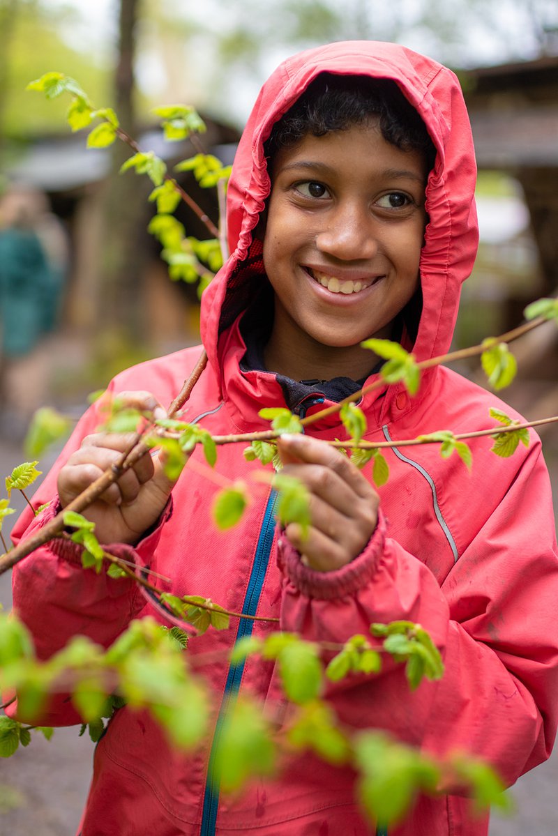 A smiling child in a raincoat stood with a tree budding with young leaves.