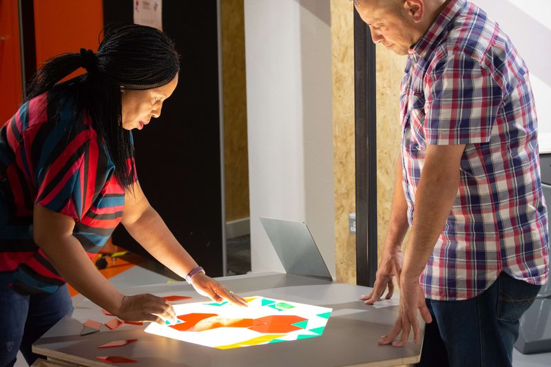 Two adults representing a mix of genders and ethnicities stand over a light table interacting with mathematical shapes