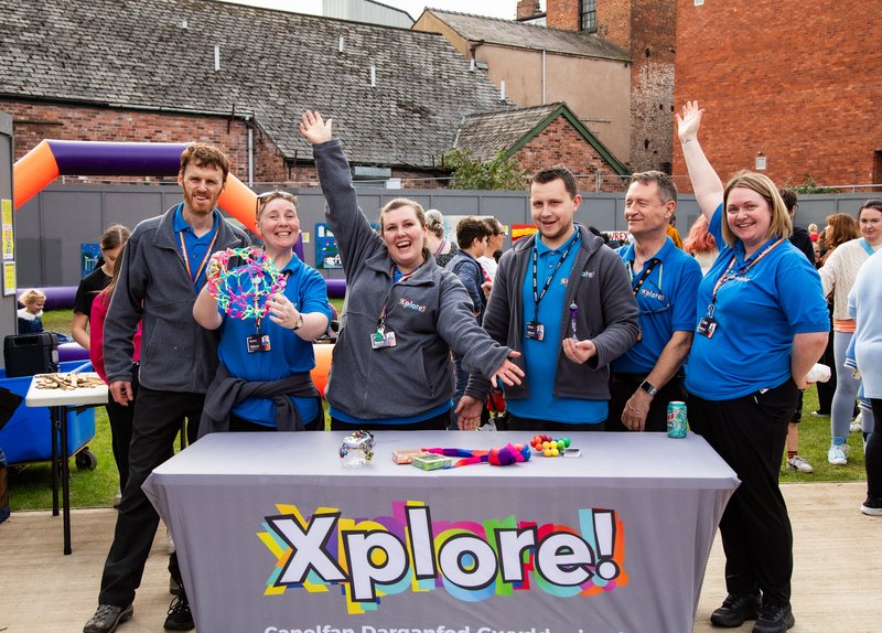 A group of six mixed gender adults stood around a table outdoors at a fair.  They are all in xplore science centre uniform and the table has a branded xplore tablecloth.