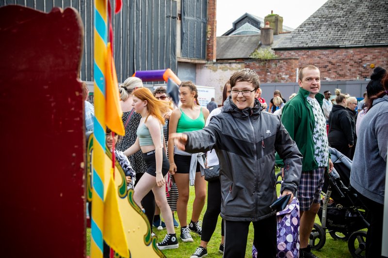 An outdoor community festival. One young person is shown smiling, in the action of throwing coming at a fairground game.